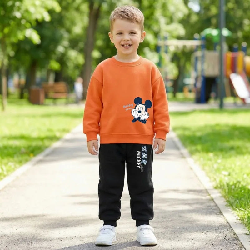A young boy stands on a paved path in a park, wearing an orange sweatshirt with a Mickey Mouse graphic and black pants with white text.