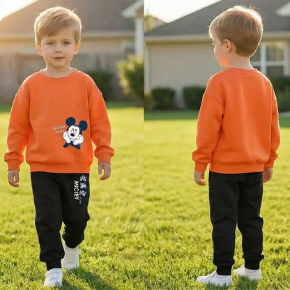 A young boy standing on a grassy lawn, shown in front and back views. He is wearing a bright orange crewneck sweatshirt with a Mickey Mouse graphic on the front, paired with black sweatpants featuring white vertical text and white sneakers.