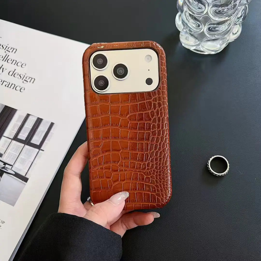 Brown crocodile-patterned phone case held by a hand on a dark surface with a magazine and glass.