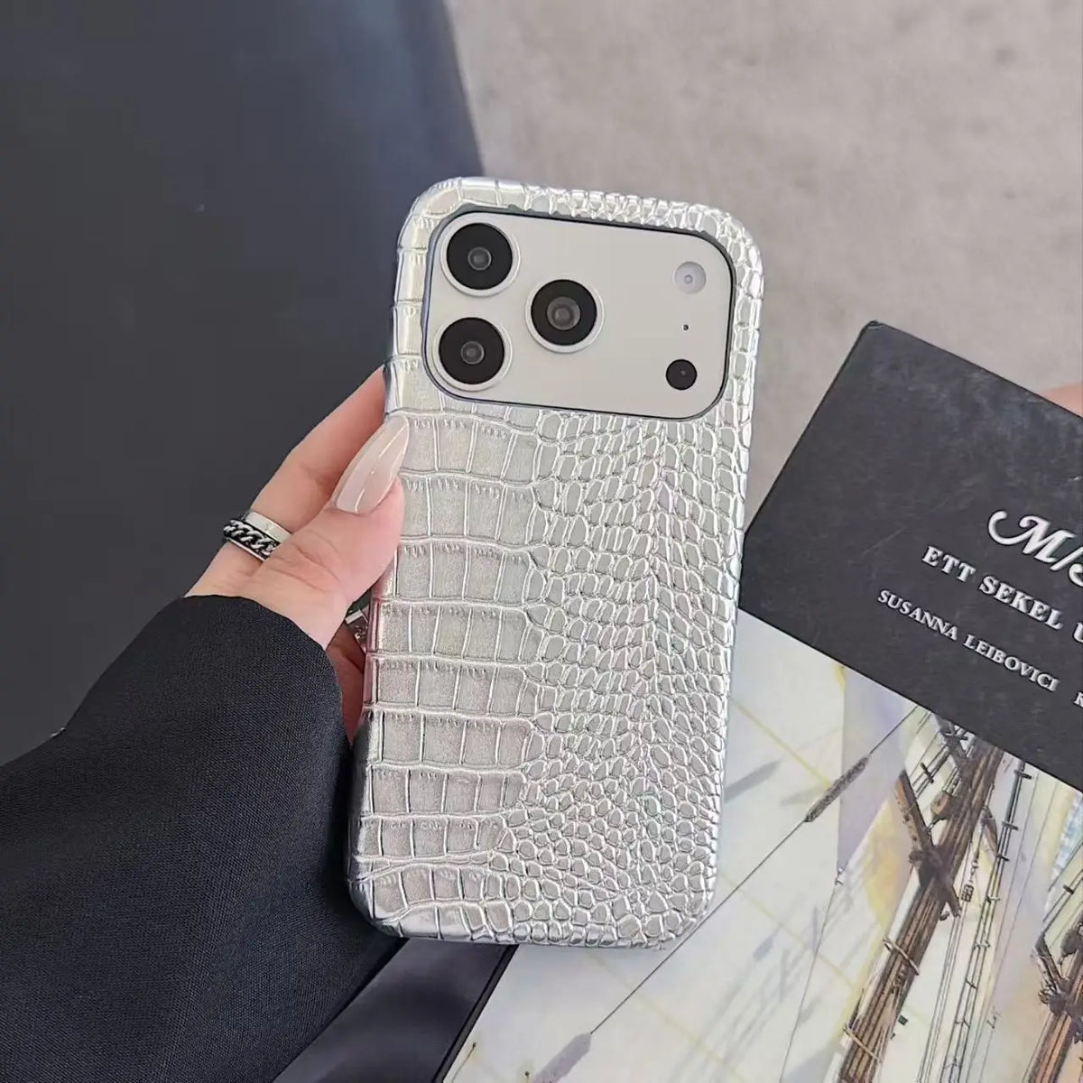A hand holding a silver crocodile-embossed phone case with a triple-camera cutout, next to a black card on a marble surface.