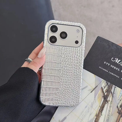 A hand holding a silver crocodile-embossed phone case with a triple-camera cutout, next to a black card on a marble surface.