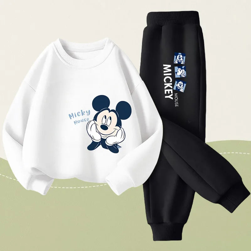 A two-piece kids' outfit set featuring a white long-sleeve sweatshirt with a Mickey Mouse graphic and black jogger pants with white Mickey text and character prints, displayed on a light beige and sage green background.