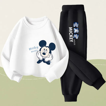 A two-piece kids' outfit set featuring a white long-sleeve sweatshirt with a Mickey Mouse graphic and black jogger pants with white Mickey text and character prints, displayed on a light beige and sage green background.