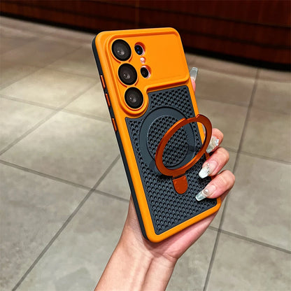 A hand holding a smartphone case with a bright orange border, dark textured center panel, and a circular brown ring stand, photographed in an indoor setting with tiled flooring.
