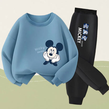 A light blue sweatshirt with a Mickey Mouse graphic and the text 'Micky Mouse' on the front, paired with black jogger pants featuring a vertical Mickey Mouse print on the leg, shown on a light beige background with a soft green curved shape at the bottom.