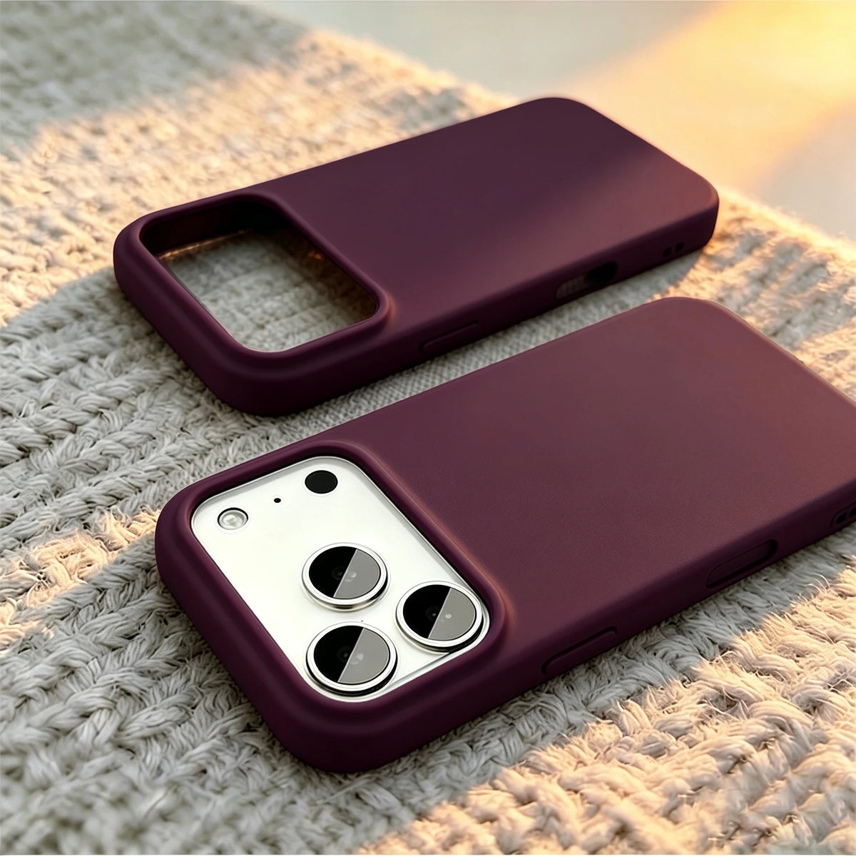 Two dark purple phone cases shown on white iPhones, resting on a textured beige knit blanket with warm sunset light.
