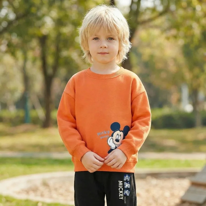 A young blond child standing outdoors in a park, wearing a bright orange long-sleeve sweatshirt with a Mickey Mouse graphic on the front and black pants with white Mickey text.