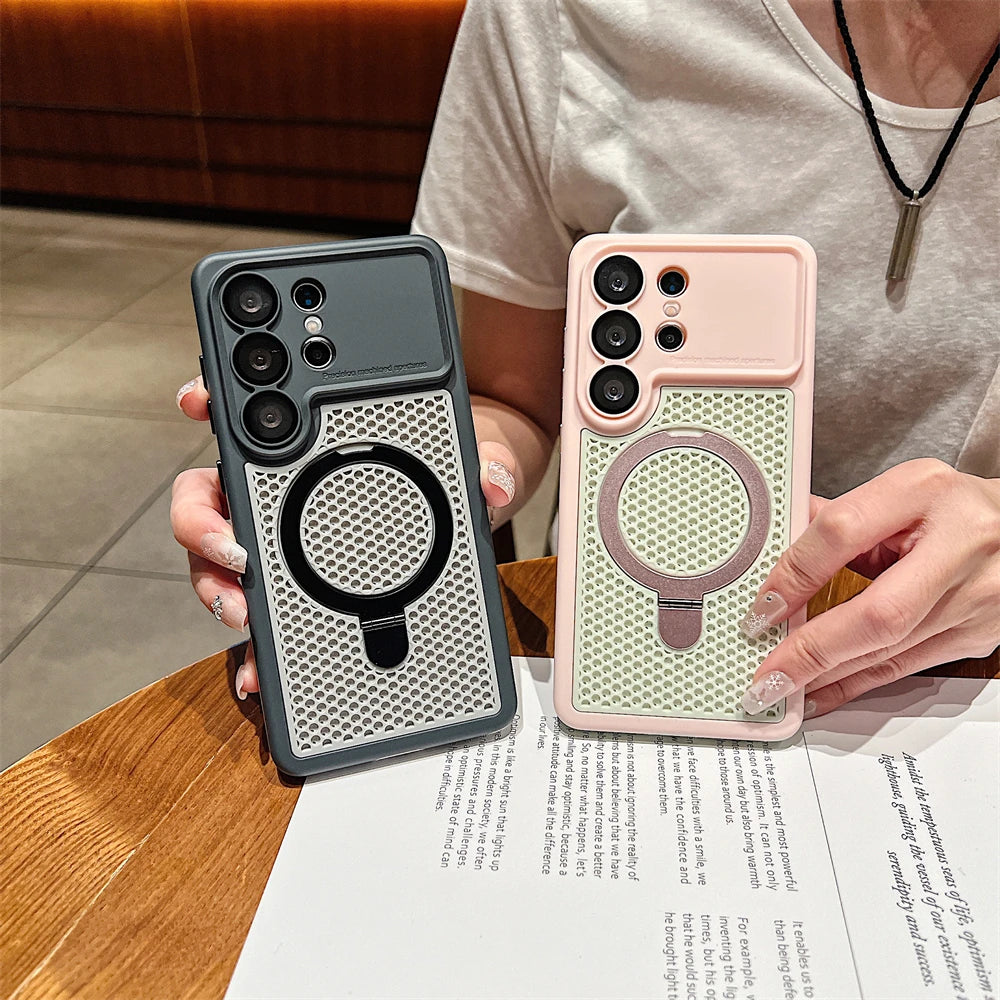 Two protective phone cases with a textured circular ring design are held by a person in a cafe setting. One case is dark gray and the other is light pink, both featuring a metallic mesh pattern and a circular MagSafe-compatible ring.