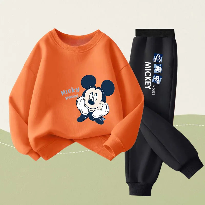 A two-piece kids' outfit set featuring an orange sweatshirt with a Mickey Mouse graphic and black jogger pants with white and blue Mickey Mouse text graphics, set against a light beige and sage green background.