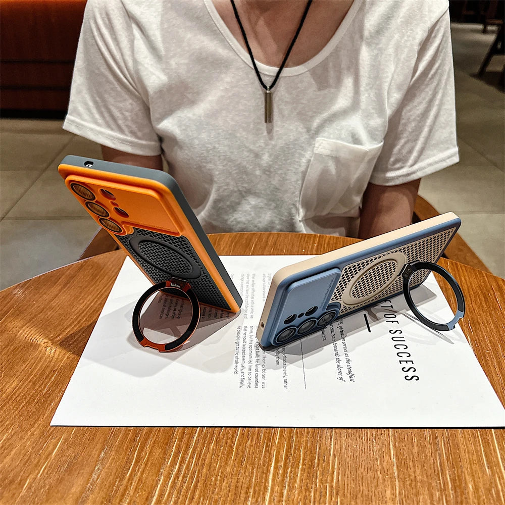 Two smartphone cases displayed on a wooden table, each holding a phone and featuring a circular ring stand. One case is orange and dark gray, and the other is light blue and beige.